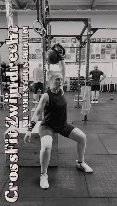 Vrouw die een kettlebell lift uitvoert terwijl ze op een box zit in een CrossFit gym.